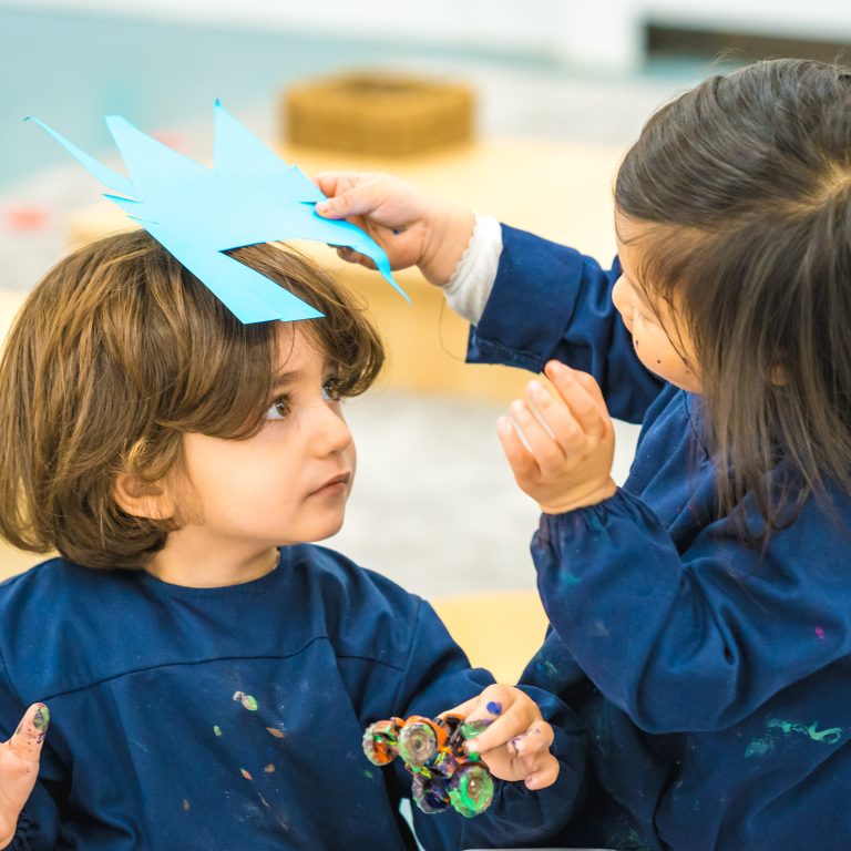 a child placing a piece of card on the top of another child's head, resembling a crown
